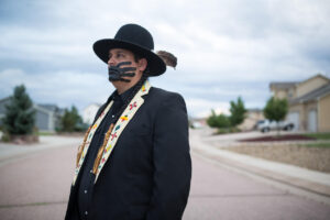 A photograph from the waist up of a Native American man standing in a suburban street, wearing a black hat with a feather and a black jacket with embroidered lapels. He has a black hand painted across his face.