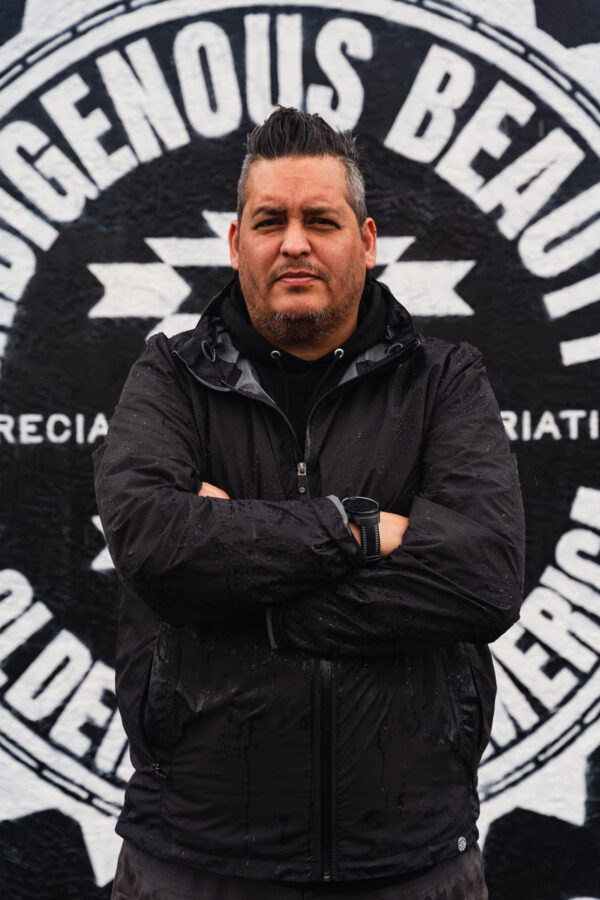 A native american man stands in front of a mural reading 'Indigenous Beauty' with his arms crossed.