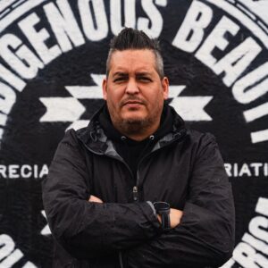 A Native American man stands in front of a mural reading 'Indigenous Beauty' with his arms crossed.